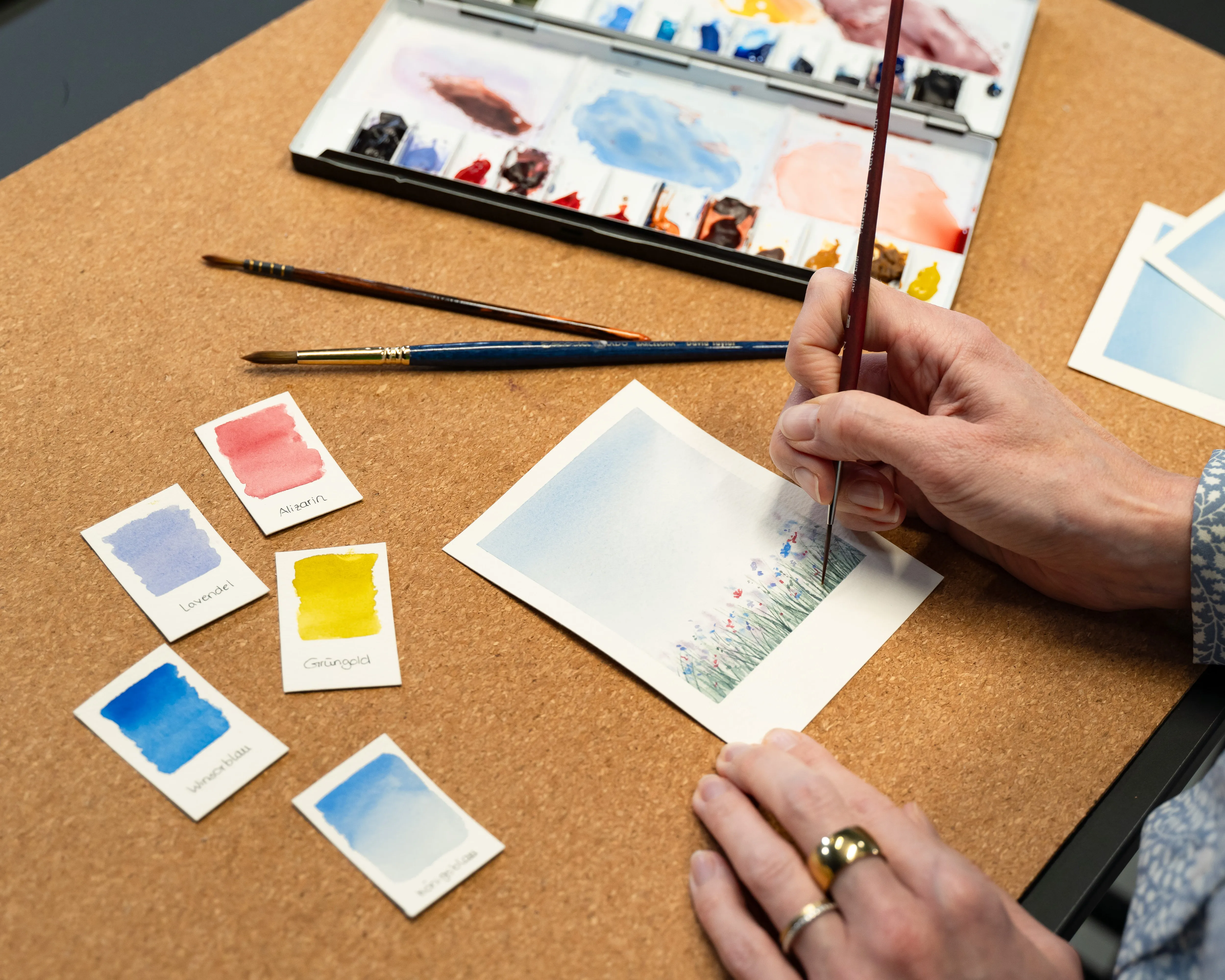 Schritt-für-Schritt-Anleitung. Eine Hand malt mit einem Pinsel auf Aquarellpapier, daneben eine Farbpalette und verschiedene kleine Aquaraell-Bilder, Bild verlinkt zum Workshop.