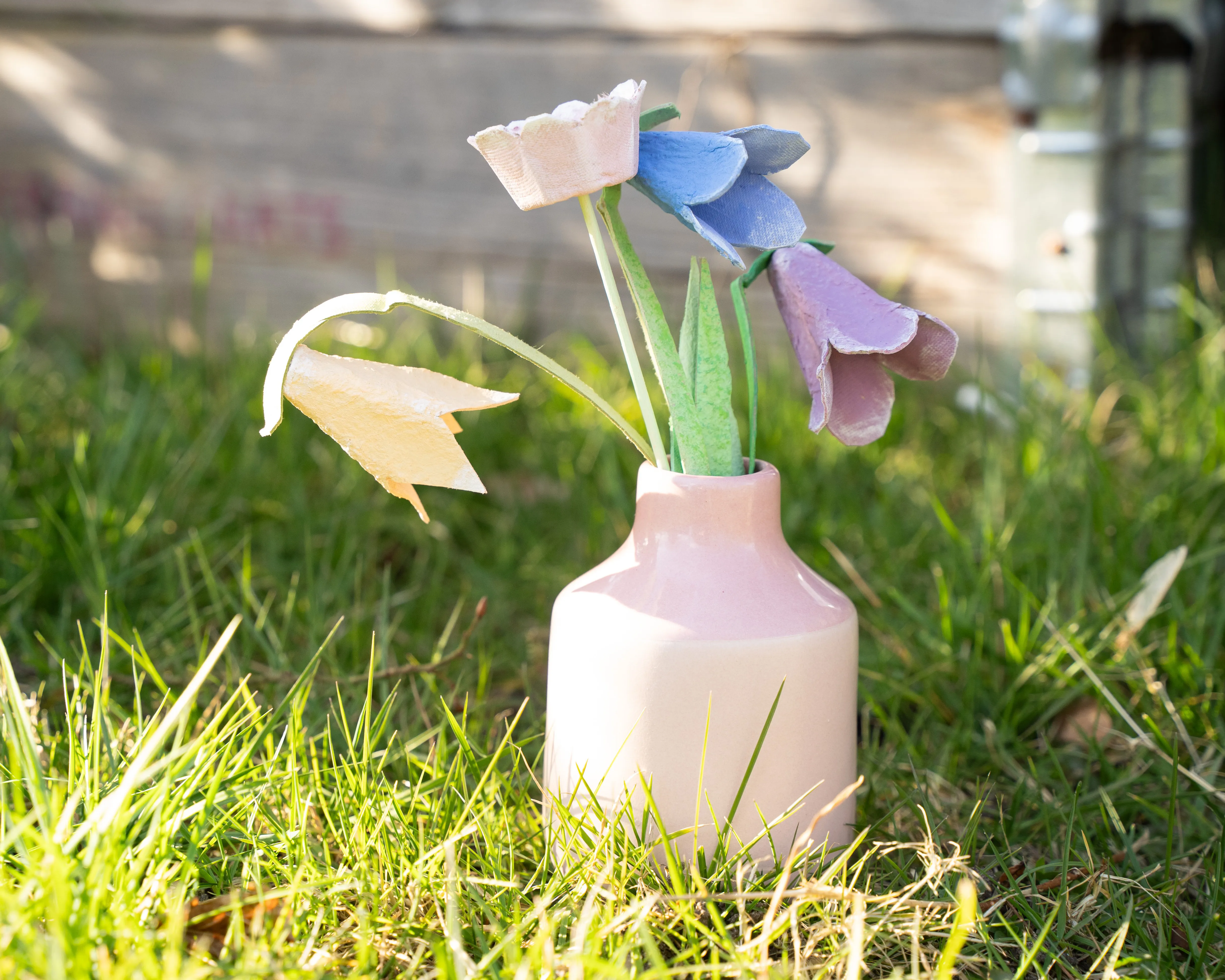 Eine kleine rosafarbene Vase steht auf einer Wiese, gefüllt mit handgemachten Blumen aus Eierschalen in verschiedenen Farben. Im Hintergrund unscharf ist eine Holzkiste zu sehen. Bild verlinkt zum Workshop.