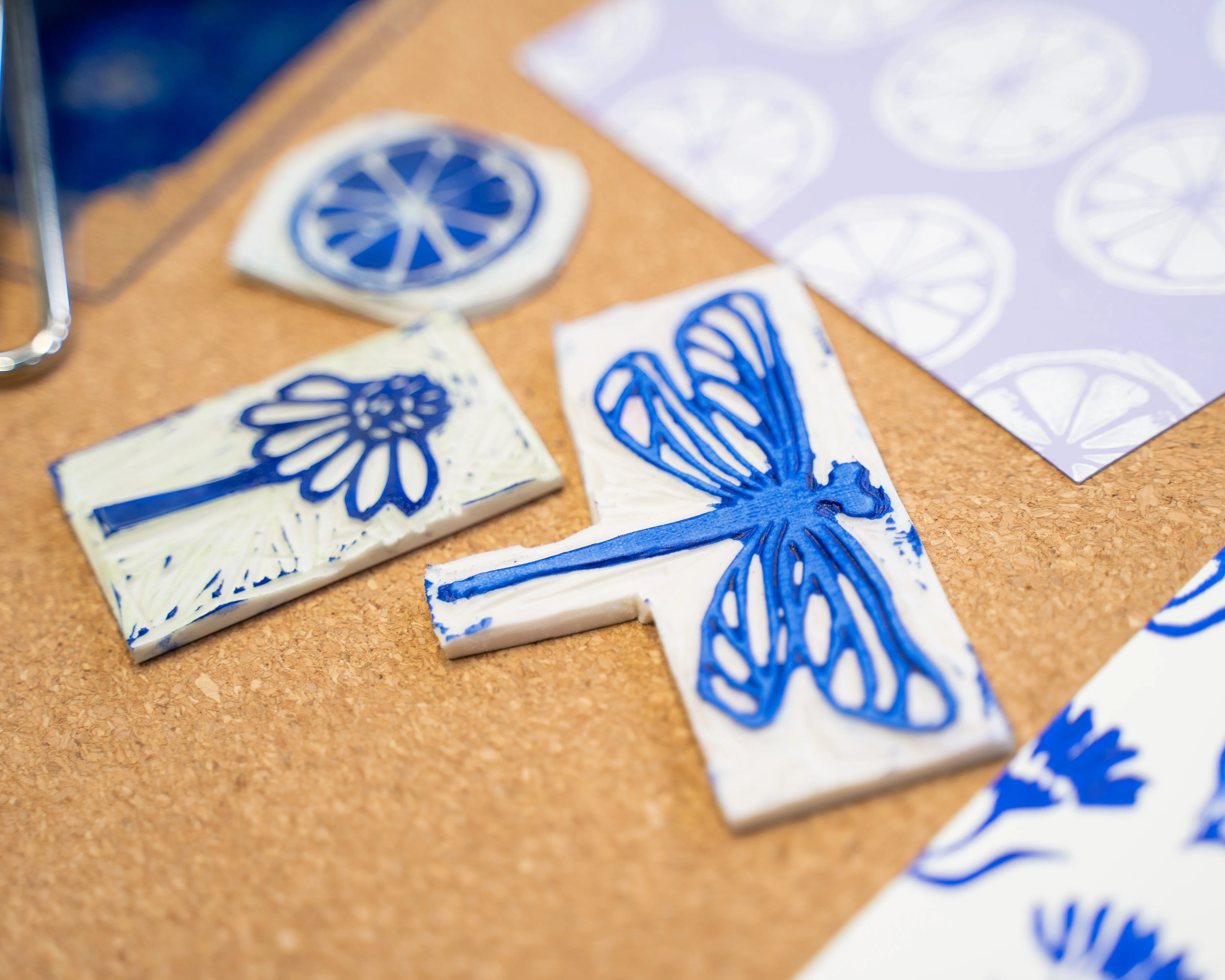 Handgeschnitzte Linoldruckstempel auf Kork, mit blauen Motiven: Libelle, Blume und Zitrusscheibe. Daneben bedrucktes Papier mit Zitronenscheibenmuster. Bild verlinkt zum Workshop.