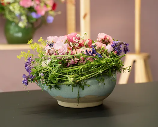 Gesteck aus lila, rosa und grünen Blumen, umgeben von einem Nest aus Zweigen, auf einem Tisch vor einer rosa Wand. Bild verlinkt zum Workshop.