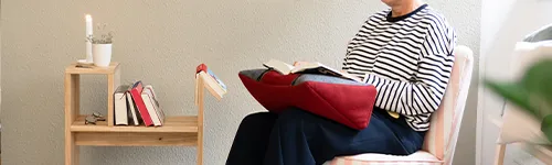 Person sitzt auf einem Stuhl und liest ein Buch auf einem roten Kissen mit Buchstütze auf den Knien. Neben ihr steht ein Holztisch mit mehreren Büchern, einer umgedrehten Brille und einer kleinen Topfpflanze.